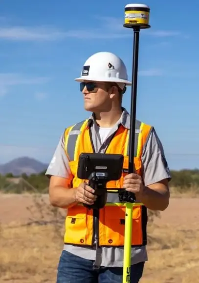 A surveyor stands outdoors in a hard hat and safety vest, holding a GPS device and pole against a clear blue sky A surveyor stands outdoors in a hard hat and safety vest, holding a GPS device and pole against a clear blue sky