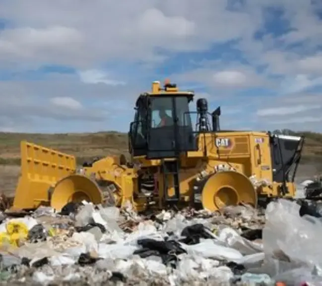 A yellow CAT bulldozer moves through a landfill, pushing aside heaps of trash under a cloudy sky A yellow CAT bulldozer moves through a landfill, pushing aside heaps of trash under a cloudy sky
